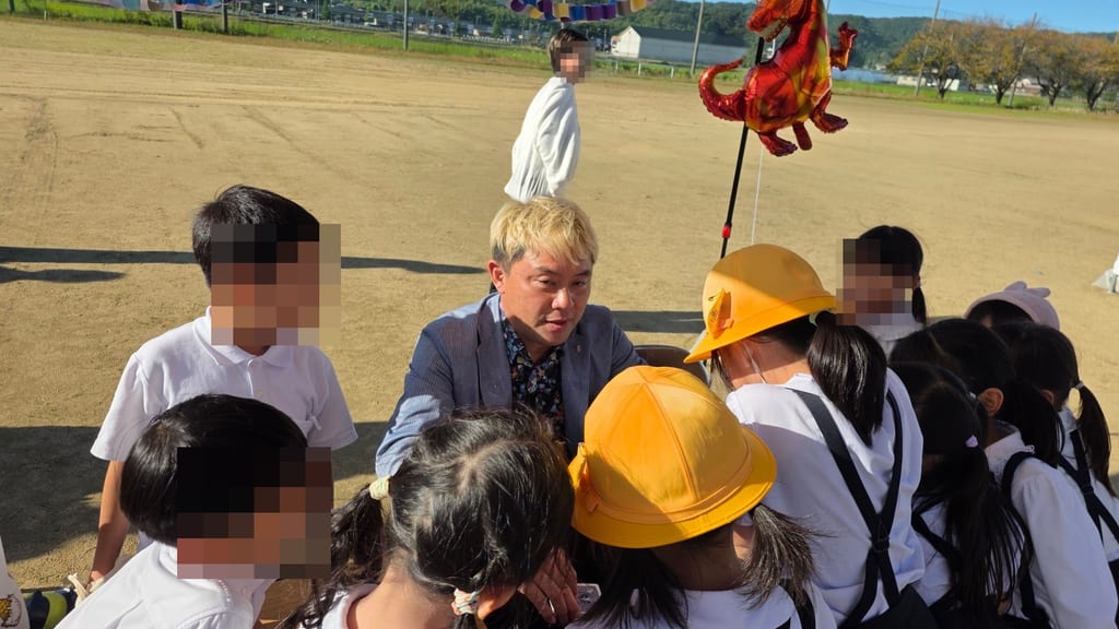 高岡市の国吉義務教育学校の校下レクリエーションで、マジック教室のテーブルを囲む黄色い帽子の子どもたちに話しかけるコンプレッサー