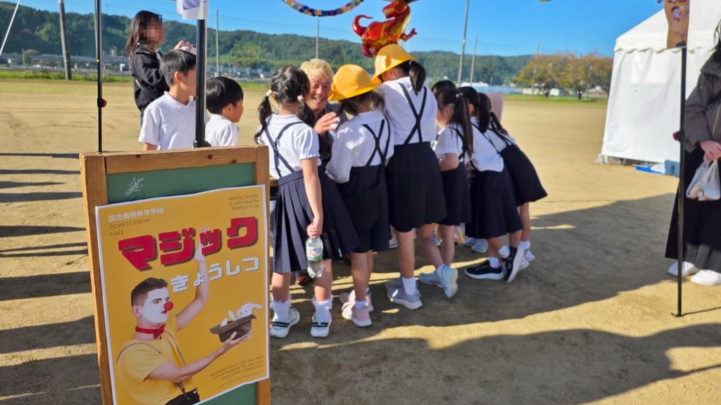 国吉義務教育学校のグラウンドに置かれた「マジックきょうしつ」の看板と、テーブルのまわりに集まる子どもたち