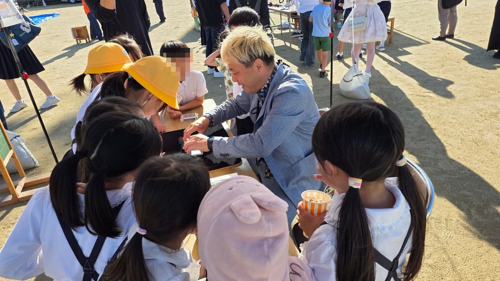 マジック教室のテーブルで、子どもたちに囲まれながらトランプのマジックを演じるコンプレッサー