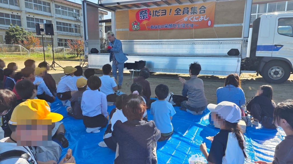 「祭りだヨ！全員集合 ＠くによし」の横断幕がかかったトラックステージの前で、ブルーシートに座る子どもたちや大人の皆さんにマジックを演じるコンプレッサー