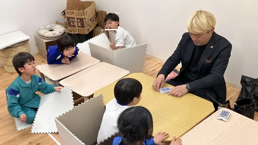 机を囲んでトランプのマジックを習う子どもたち