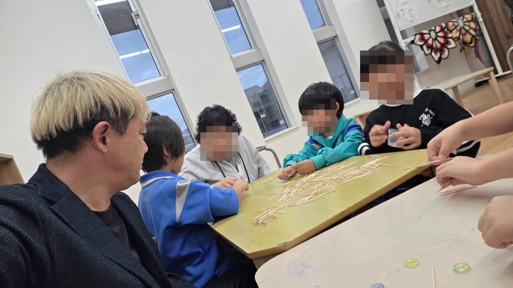 高岡シュタイナー学童保育で、楊枝が広がった机を子どもたちと囲むマジシャン・コンプレッサー。手前の机にはおはじきが置いてある