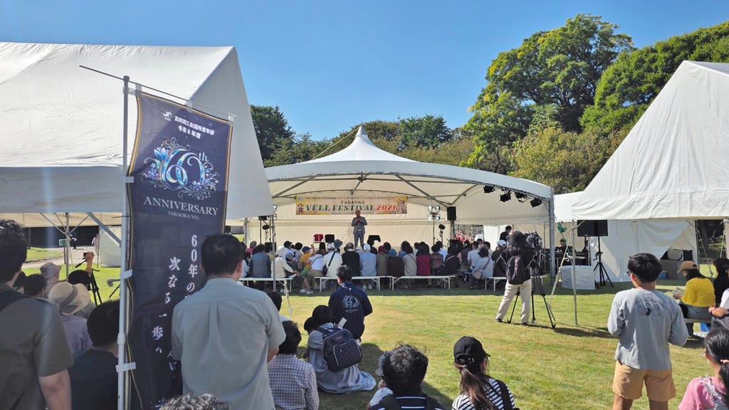 雲ひとつない青空の下、高岡古城公園の芝生に立つ白いテントのステージと、ステージに立つマジシャン・コンプレッサーを見る大勢のお客さま