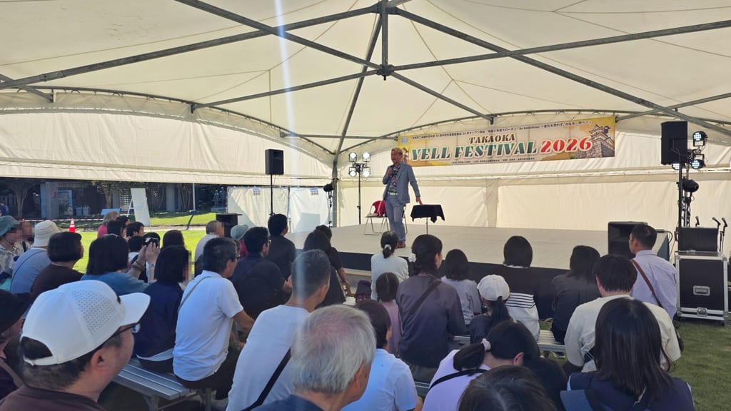 高岡古城公園のTAKAOKA YELL FESTIVAL 2026で、テントのステージに立ってマイクで話すマジシャン・コンプレッサーと、ベンチに座るお客さま
