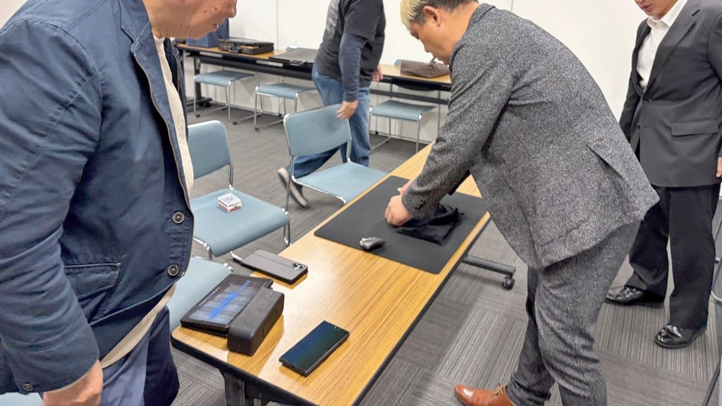 まじっぱで、黒いマットの上に置いた布の袋を整えるマジシャン・コンプレッサーと、そばで見守るメンバー