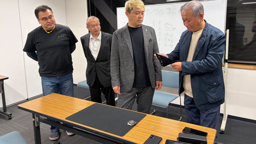 まじっぱで、ホワイトボードの前に並んだメンバーのひとりが、黒い袋からコインを1枚引く様子。隣にはマジシャン・コンプレッサー