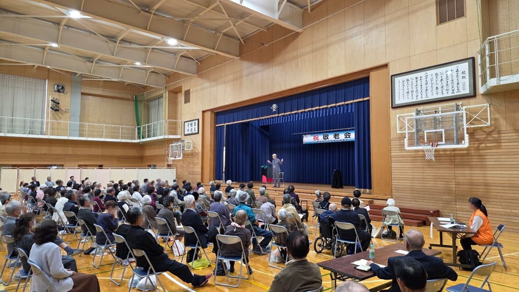 豊田小学校の体育館のステージに「祝 敬老会」の横断幕がかかり、手前に並べたパイプ椅子の客席からステージのコンプレッサーを見ている様子
