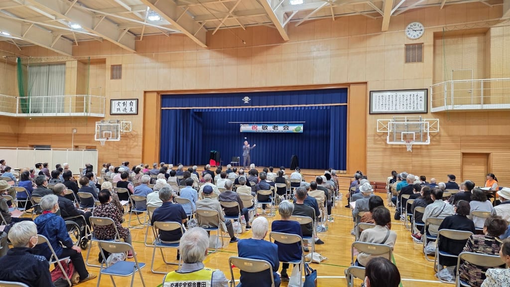 豊田小学校の体育館の後方から見た敬老会の客席と、「祝 敬老会」の横断幕の下で両手を広げるステージ上のコンプレッサー