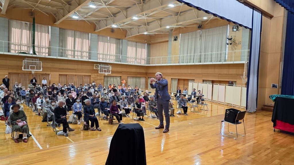 富山市の豊田小学校の体育館で、敬老会の客席に向かって両手を顔の前に構えるコンプレッサー