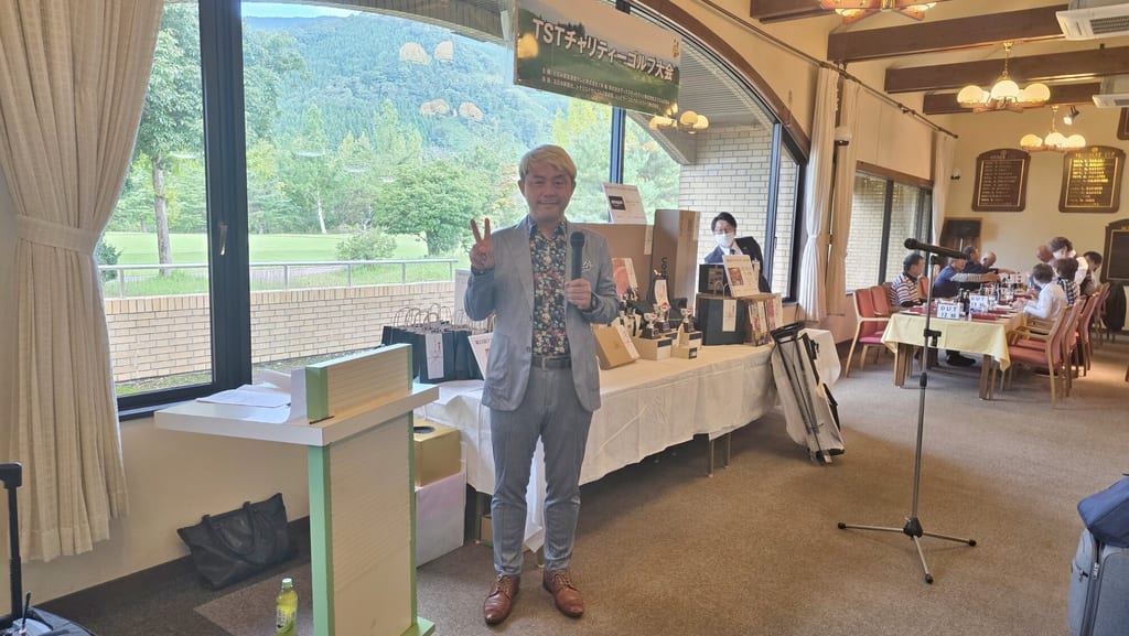 南砺市のトナミロイヤルゴルフ倶楽部のレストランで、TSTチャリティーゴルフ大会の横断幕と賞品が並ぶテーブルの前に立ち、マイクを持ってピースをするコンプレッサー